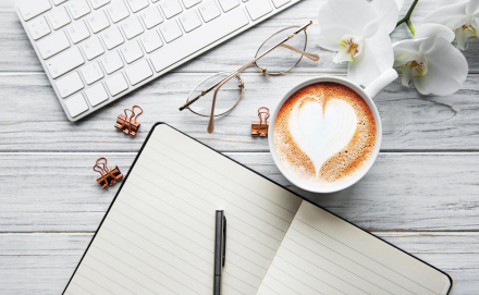 A cup of coffee with heart pattern on a table with a notepad and computer keyboard